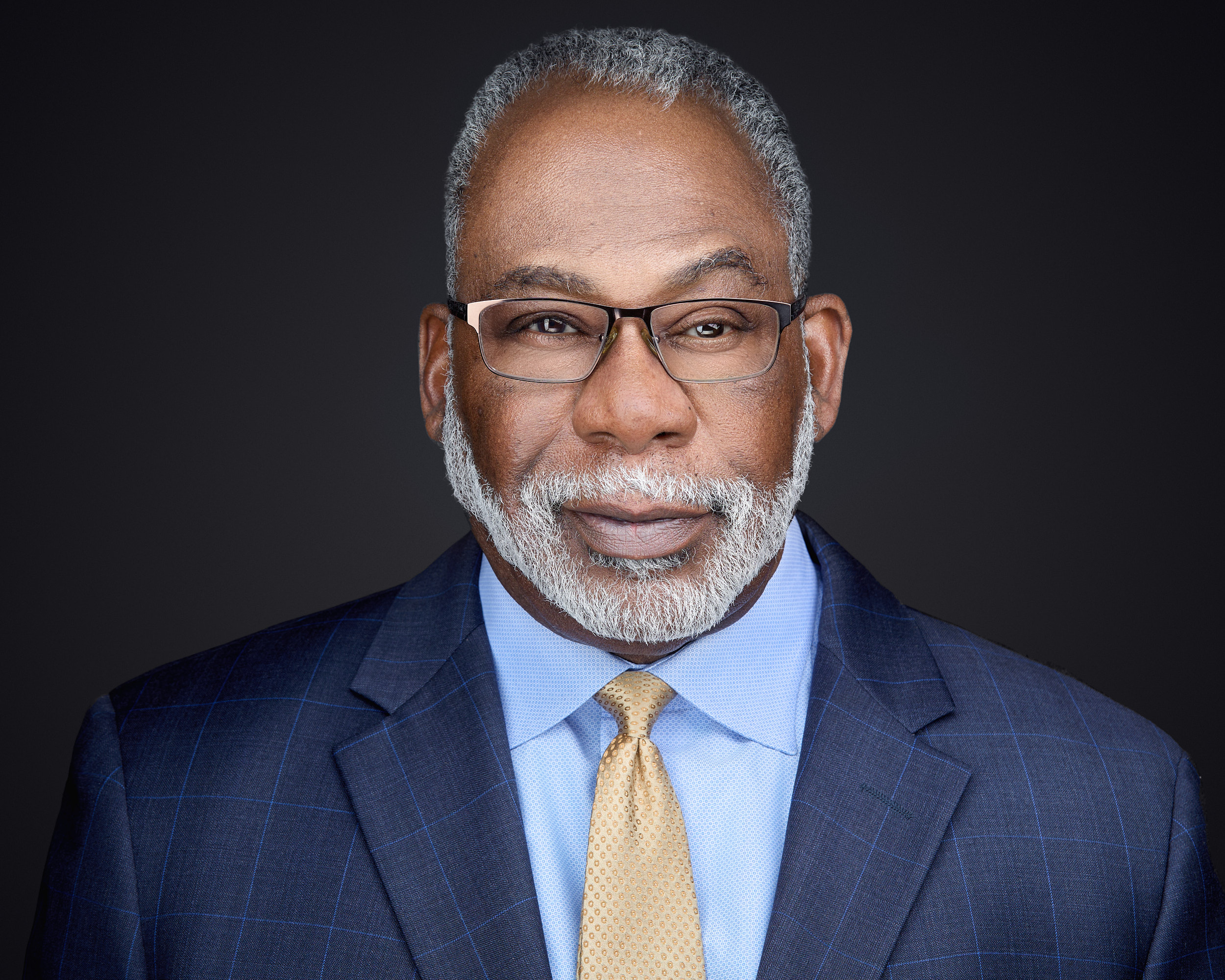 Alt Text Studio headshot of a smiling, older Black male executive wearing glasses and a dark blue suit jacket with a light blue shirt.