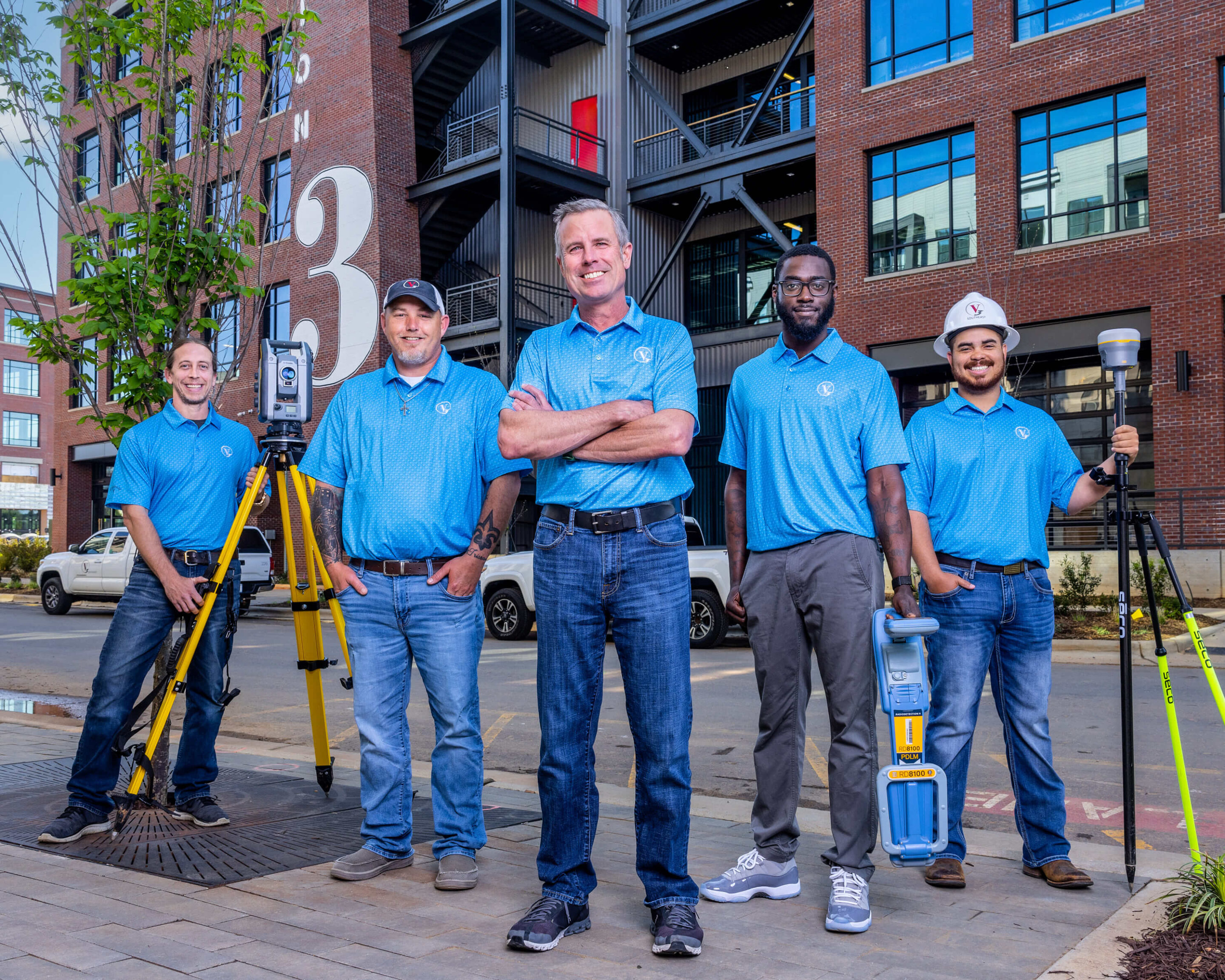 V3 Surveying Team Group Portrait – Charlotte Commercial Photography Alt Text Outdoor group portrait of five V3 Surveying team members wearing matching blue shirts and high-visibility safety vests, posing together with surveying equipment on location in Charlotte, NC.